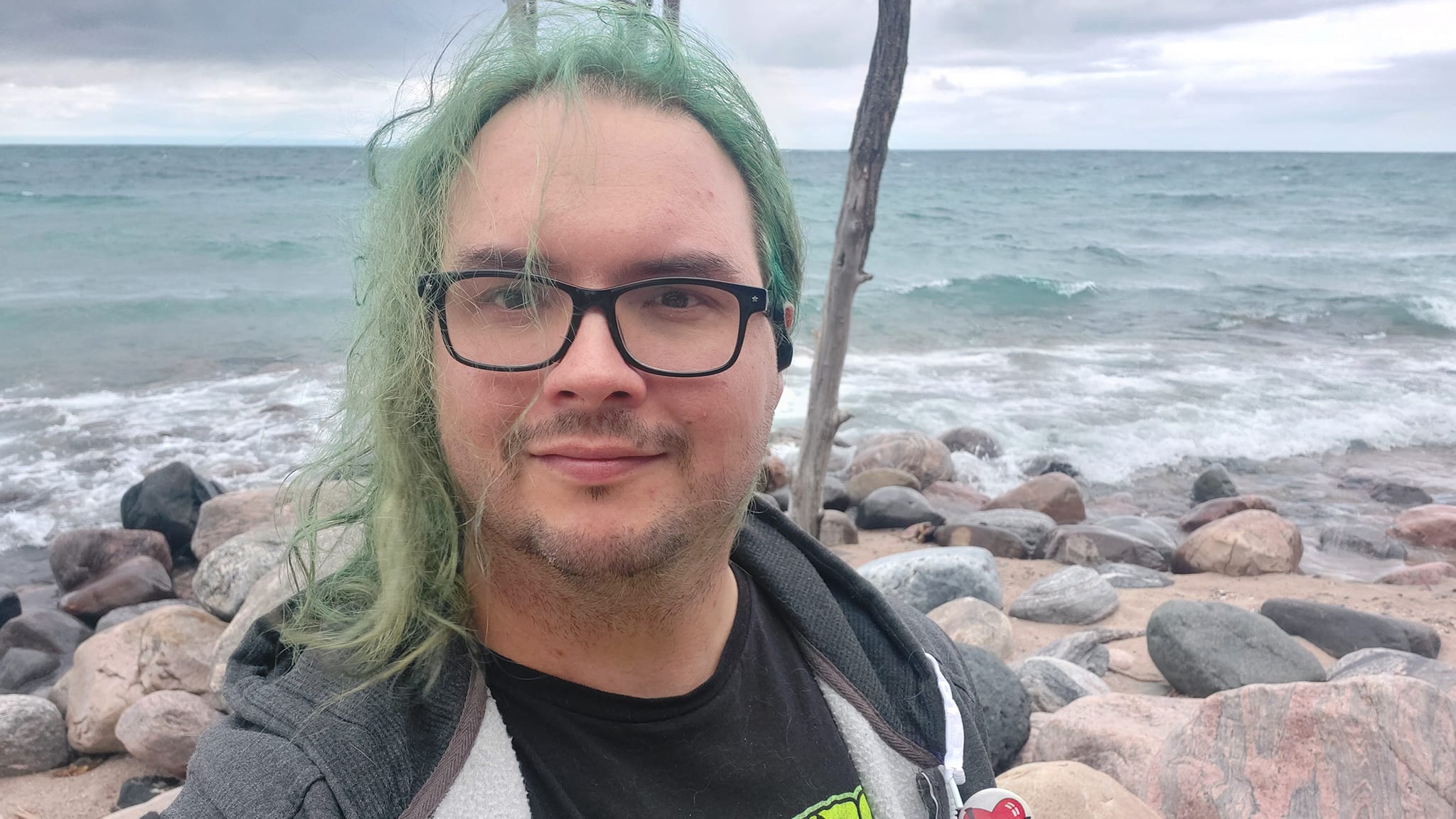 A man with long green hair and no beard stands in front of Lake Huron, waves crashing on the breakwater, Photo 2